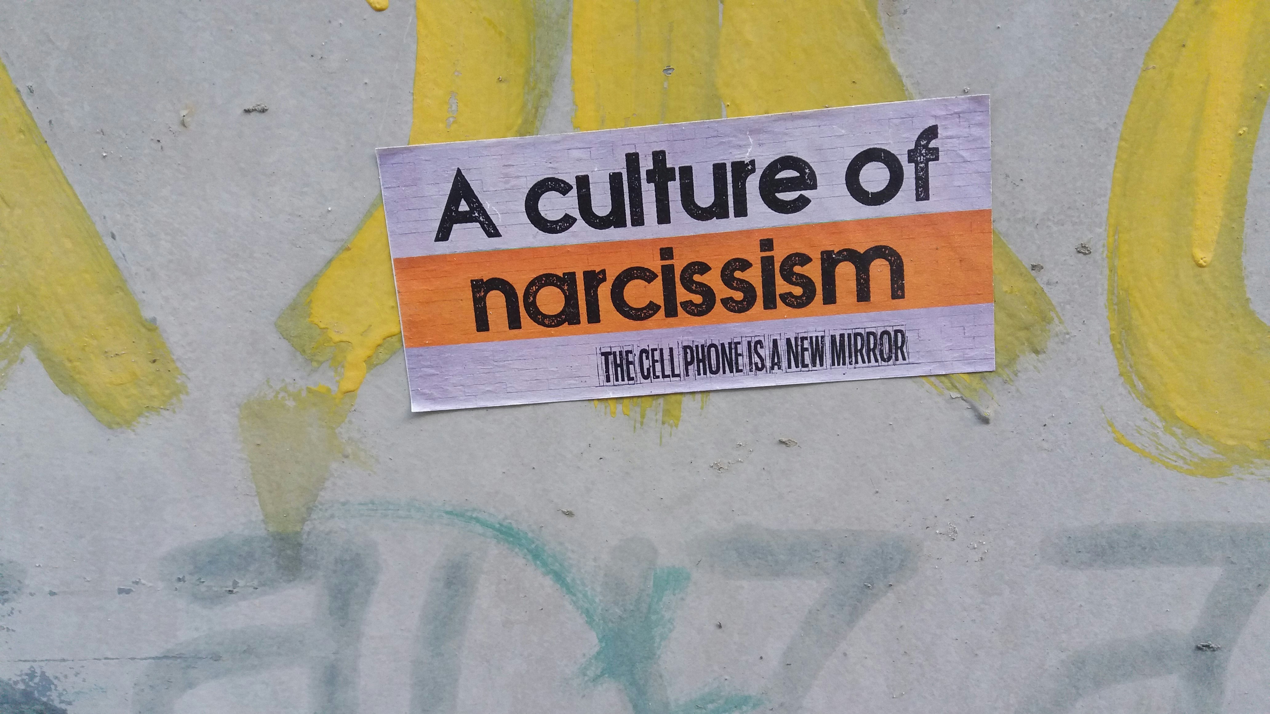A paper bill glued to a wall of graffiti that says "A CULTURE OF NARCISSISM" on the top and "The phone is the new mirror" below.